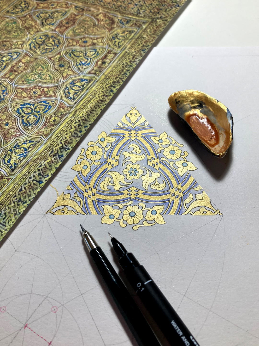 Decorative illuminated pattern in gold and blue on paper with a pen, pencil and shell of gold.