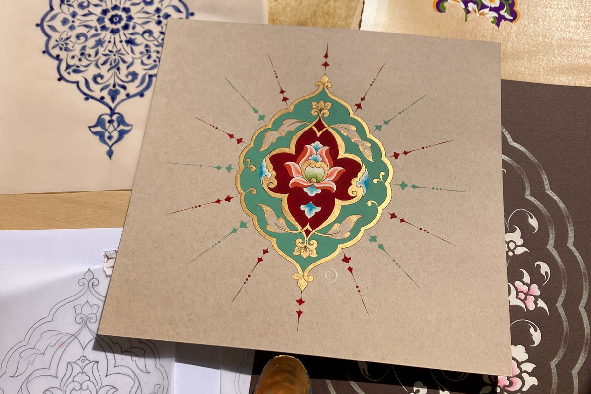 A square toned card painted with a shamsa motif design in gold, red and malachite, laid on top of other painted artworks.